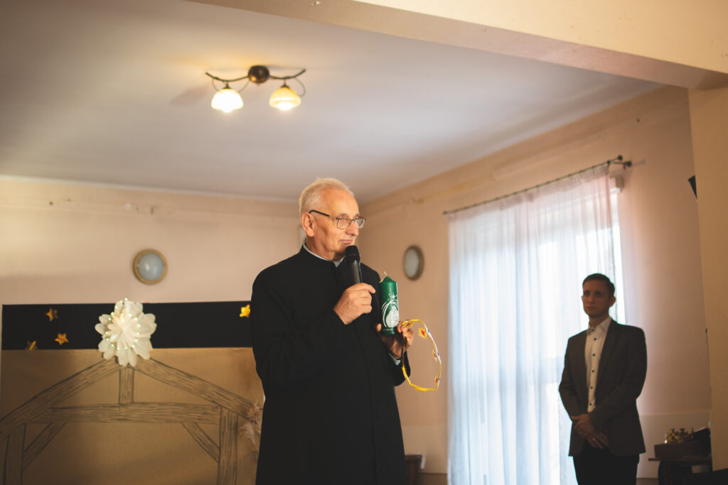 Ksiądz Proboszcz Zdzisław Balon stoi na scenie i składa świąteczne życzenia a w ręku trzyma świece. 
