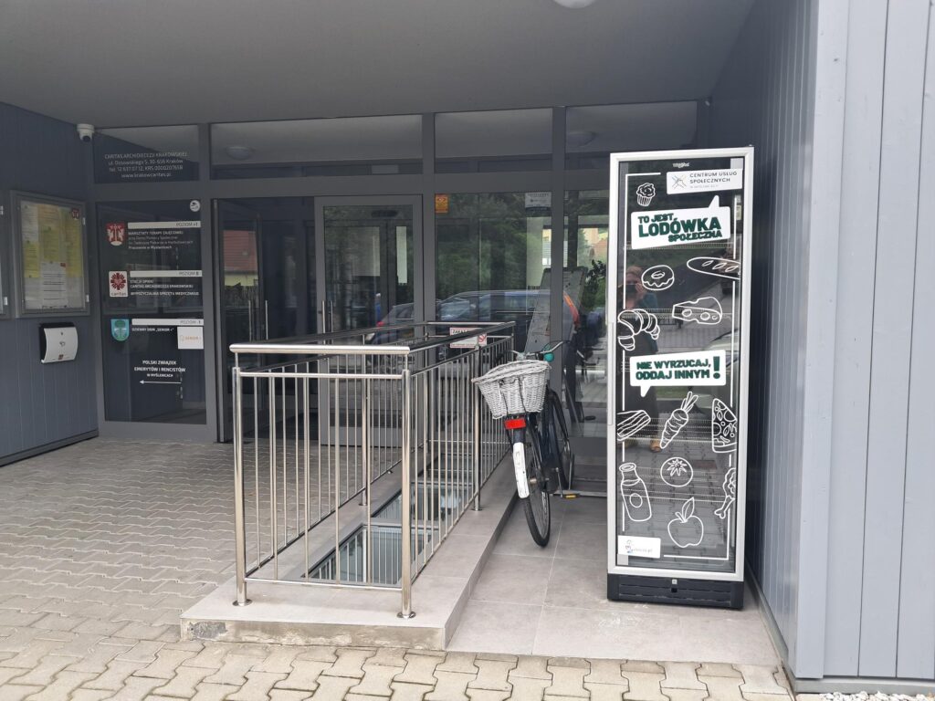 Po prawej stronie widać lodówkę społeczną na tle budynku Caritas przy ul. Średniawskiego 35